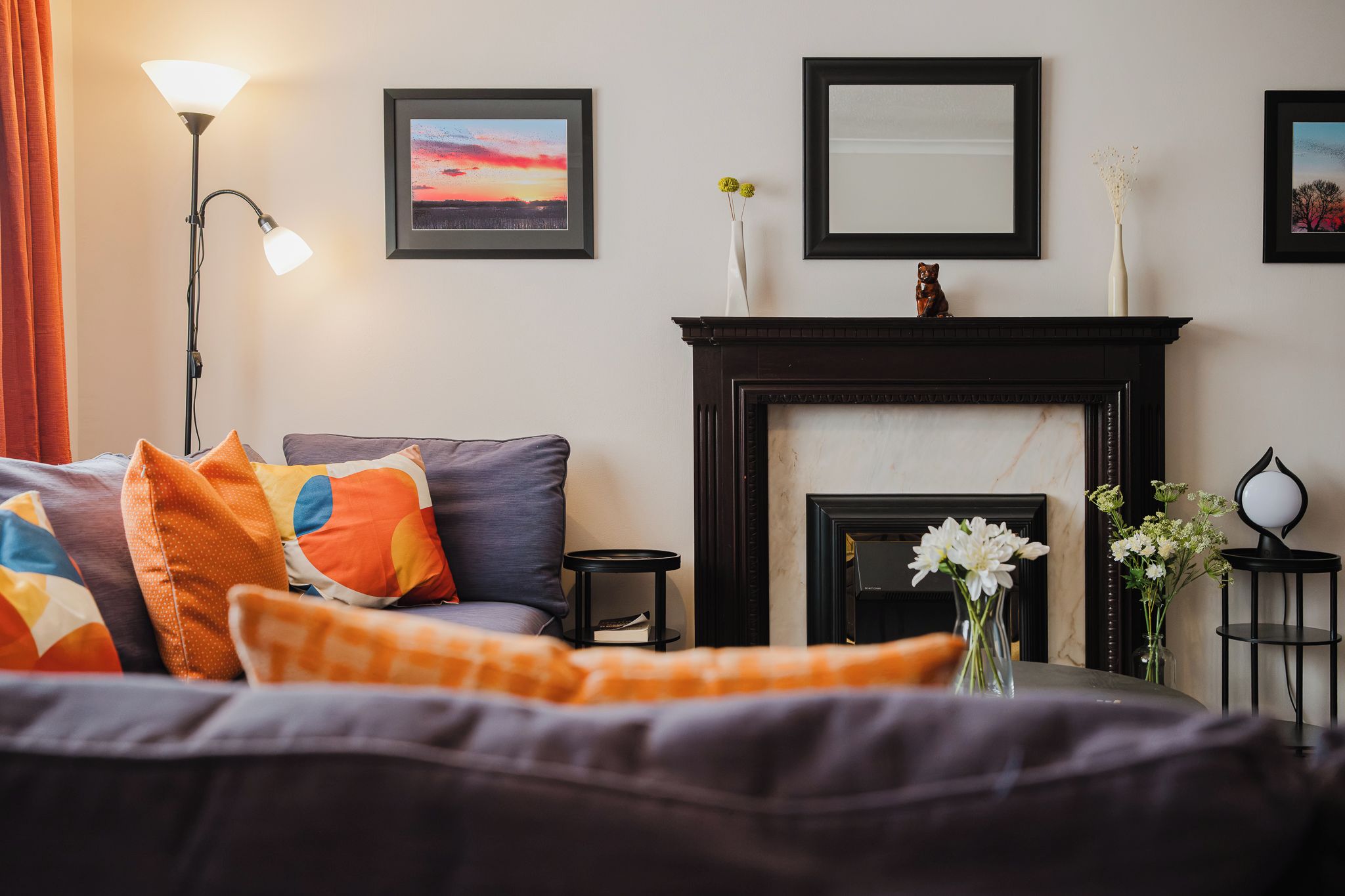 A living room with a fireplace, a sofa with colorful cushions, a lamp, and framed artwork on the wall.