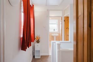 A narrow hallway with a white door on the right, a small white cabinet with a vase of flowers, and a window with a view of a bathroom. - Tor View-Stunning Home near Glastonbury (Street)