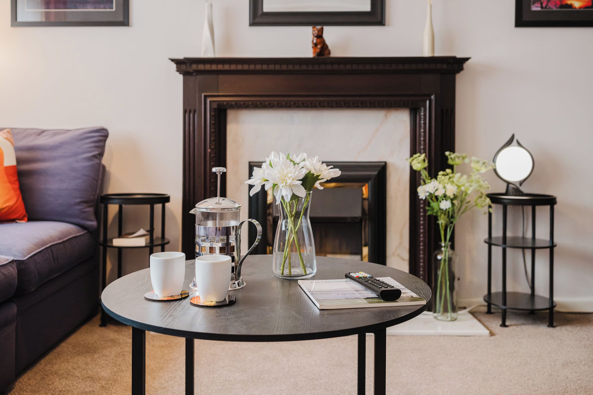 A round table with a coffee maker, two white cups, a remote control, and a book.