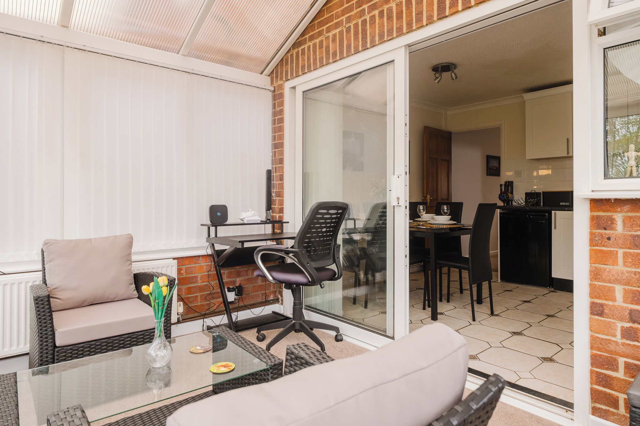 A patio area with a glass table, two chairs, a small table with a vase of flowers, and a brick wall.