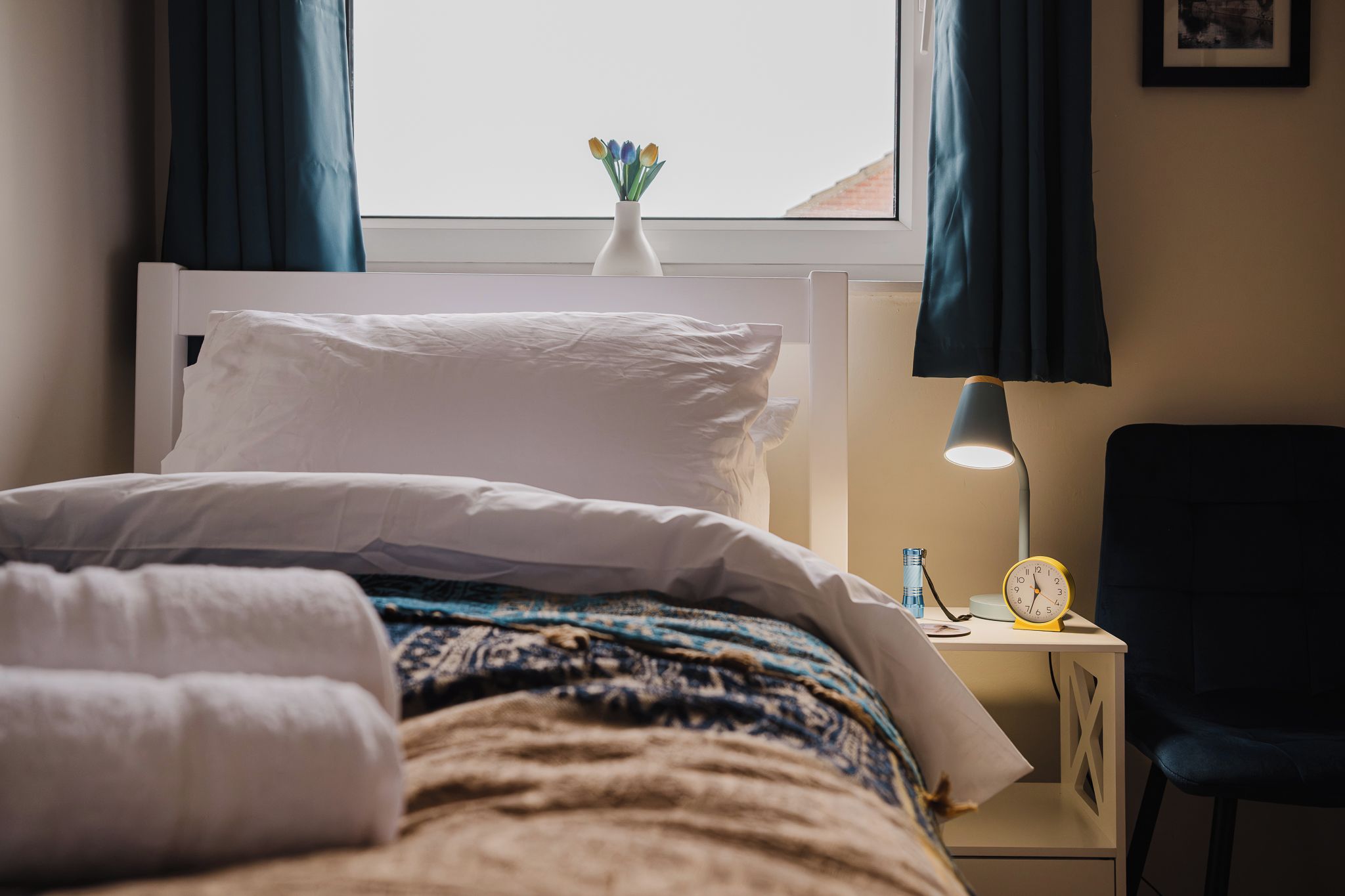 Una cama bien hecha con ropa blanca, una mesita de noche con lámpara, un reloj y un pequeño florero con flores.