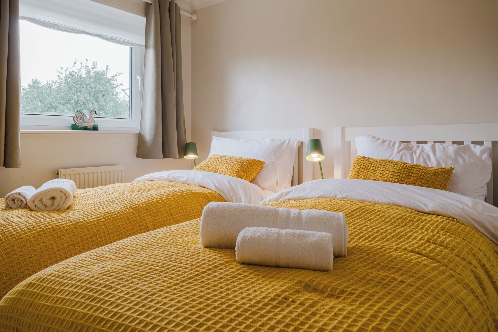 A hotel room with two beds, each with yellow bedding and white pillows, a window with curtains, and a radiator.