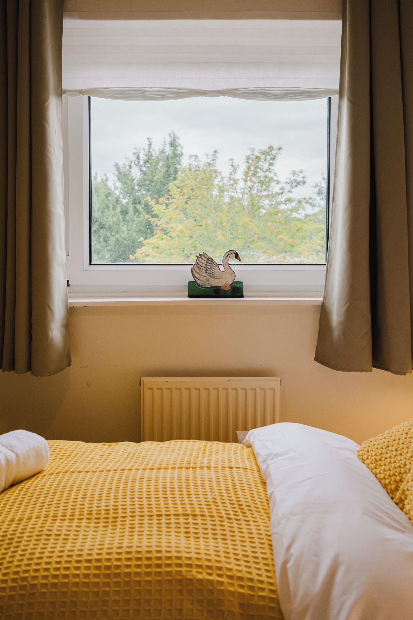 A room with two beds, a radiator, a window with curtains, and a swan decoration on the windowsill.