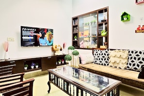 A living room with a sofa, a coffee table, a TV mounted on the wall, and a bookshelf with decorative items. - Nivera Stays 4BHK Ground Floor Home near Fortis, IGI Airport, South Delhi (New Delhi)