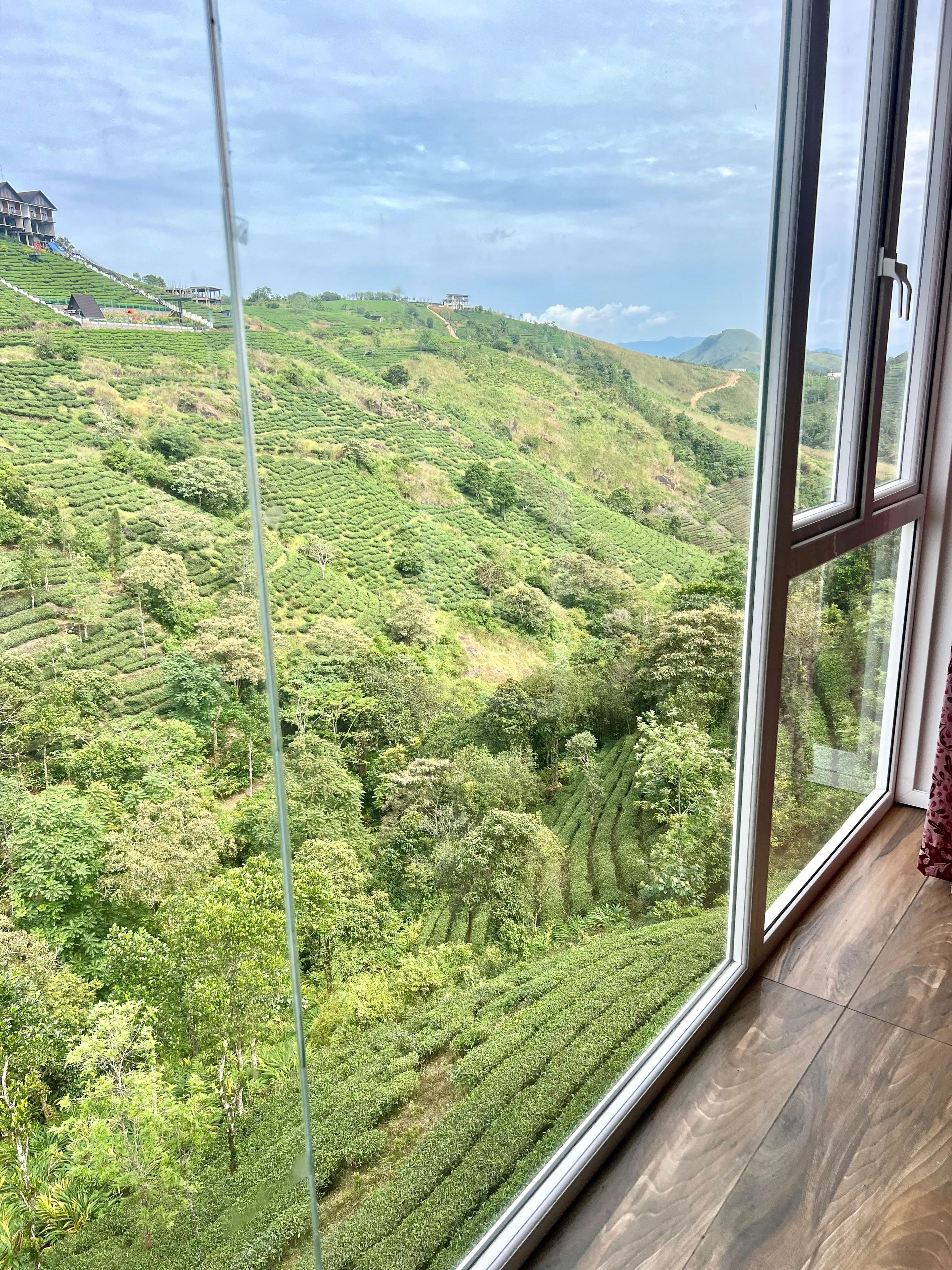 Vista a través de una ventana de una habitación con un amplio ventanal que da a un exuberante paisaje verde con campos en terrazas y colinas al fondo.