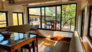 A room with a glass dining table, wooden chairs, a cushioned bench, and large windows offering a view of the outdoors.