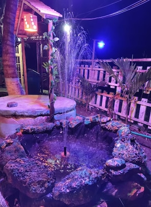Una fuente rodeada de rocas y plantas, bajo una estructura de madera con un letrero.