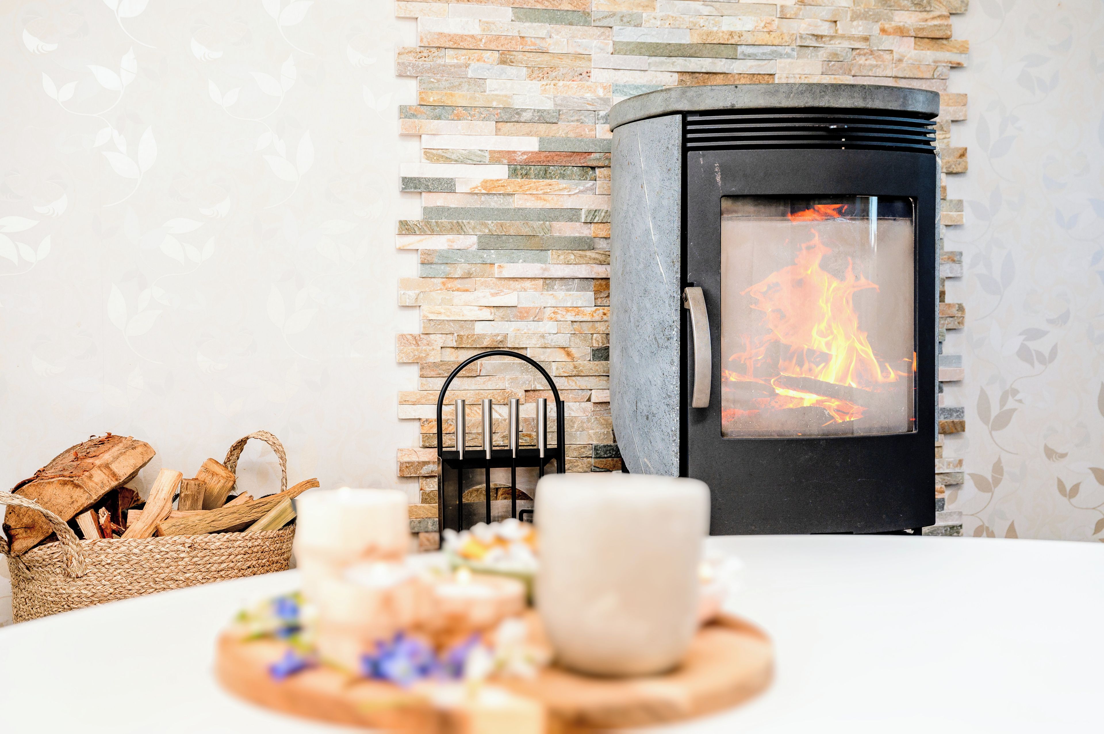 A cozy room with a stone wall, a wood-burning stove, a woven basket with firewood, and a candle on a table.