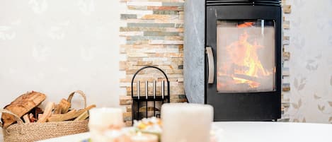 A cozy room with a stone wall, a wood-burning stove, a woven basket with firewood, and a candle on a table.