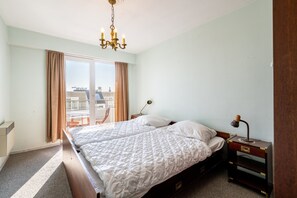 A hotel room with a large bed, a bedside table with a lamp, and a view of the city through the window. - Apartment in Blankenberge by the Beach (Blankenberge)