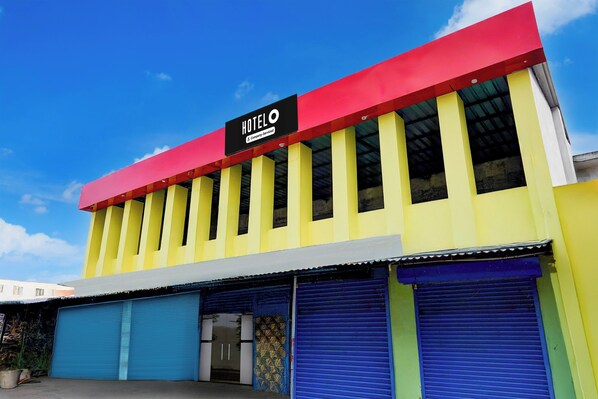 A hotel with a red and yellow facade, a black sign with 'HOTEL' and 'Company-Serviced' text, and blue roller shutters on the ground floor. - Hotel O Poigai Near Airport (Chennai)