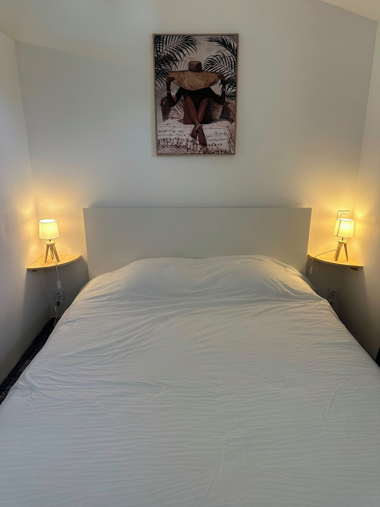 Ein Bett mit weißer Bettdecke, begleitet von zwei Nachttischlampen auf Holzständern, und einem Wandbild darüber.
