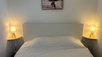 Ein Bett mit weißer Bettdecke, begleitet von zwei Nachttischlampen auf Holzständern, und einem Wandbild darüber.