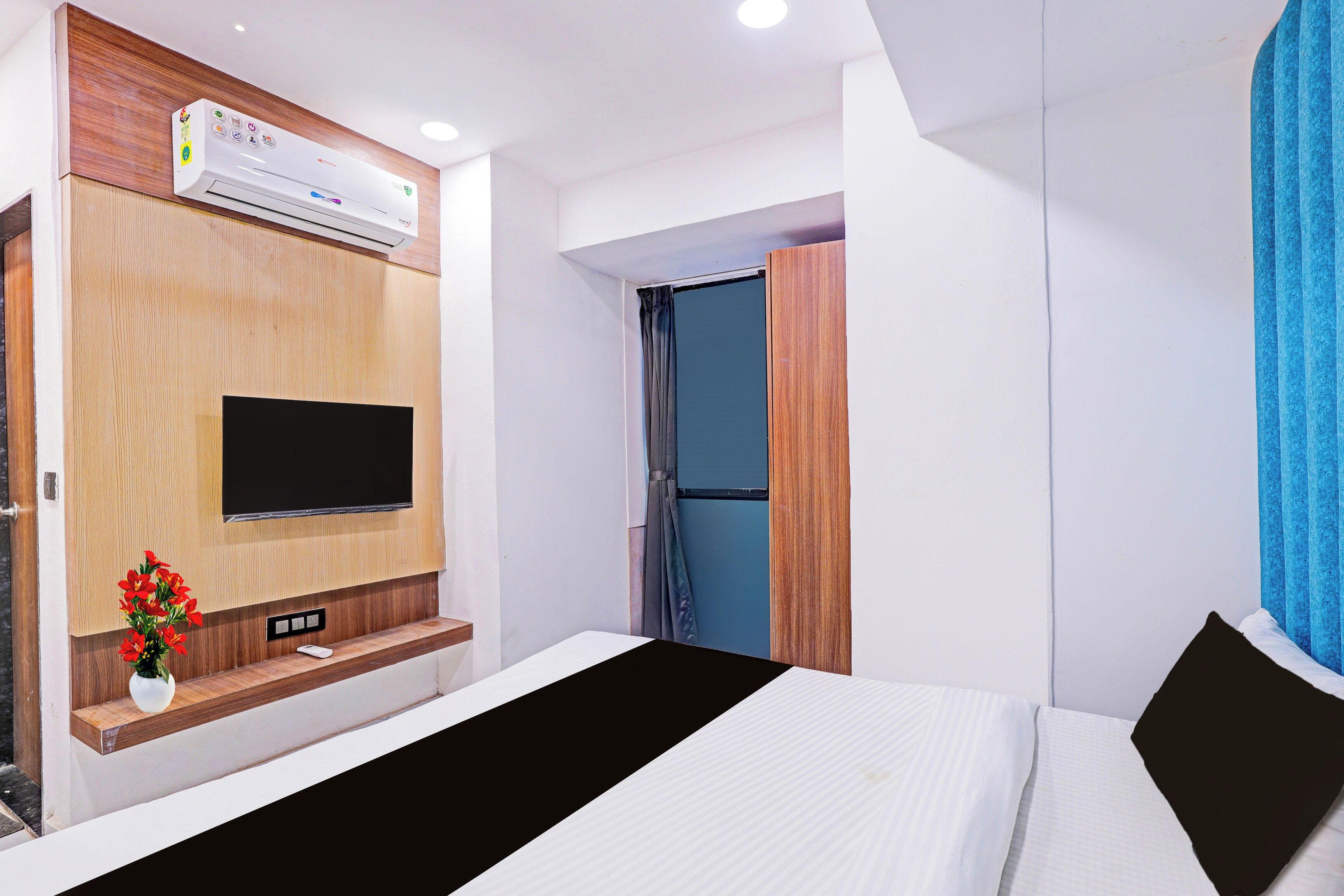 Ein Hotelzimmer mit einem Bett, einem an der Wand montierten Fernseher, einer Klimaanlage und einer kleinen Ablage mit einer Blumenvase.