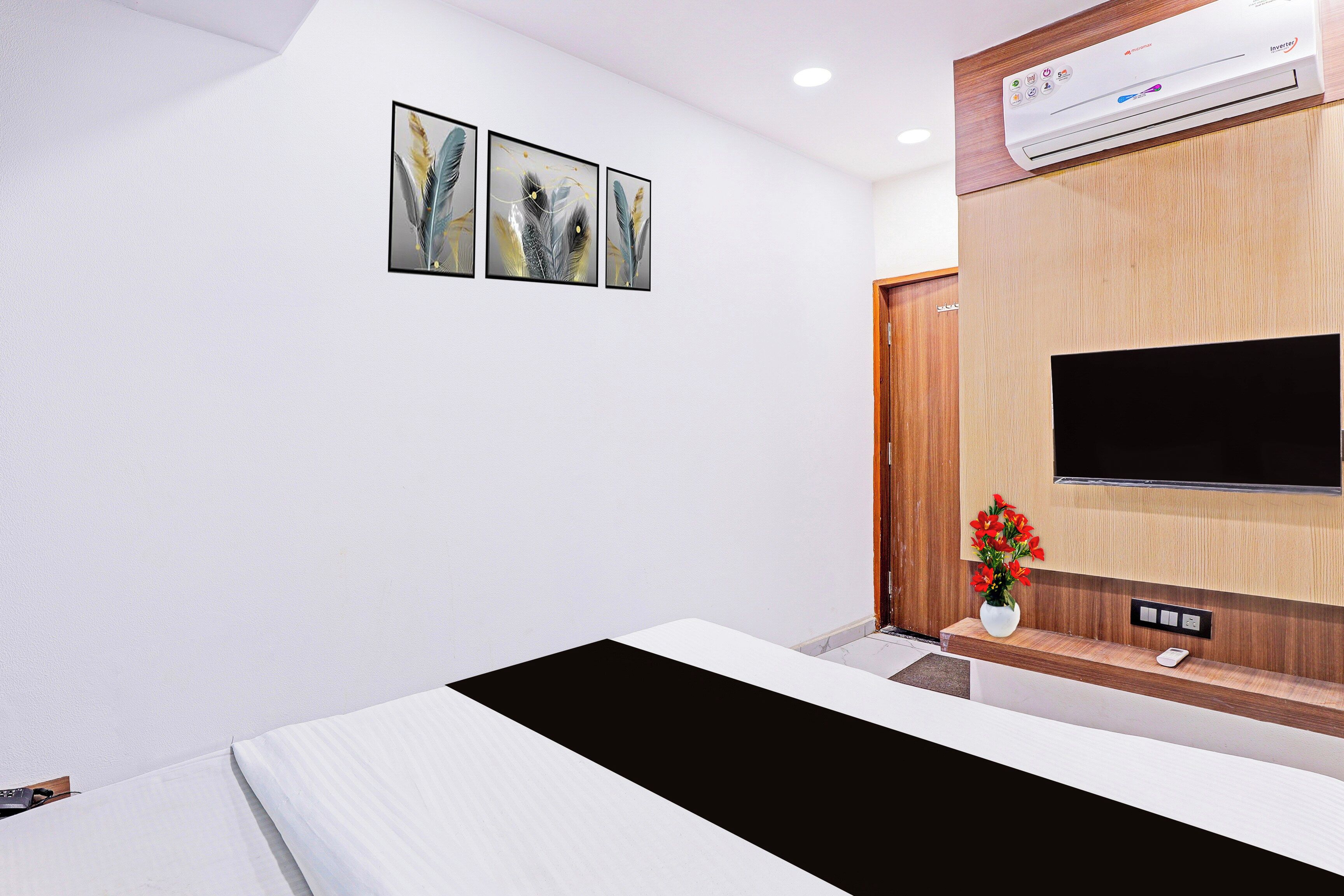 Une chambre d’hôtel comprenant un lit, un téléviseur à écran plat fixé sur un panneau en bois, une petite table avec un vase de fleurs et trois œuvres d’art encadrées accrochées au mur.