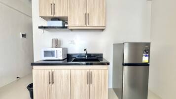 A compact kitchen with light wood cabinets, a stainless steel refrigerator, a microwave, and a black countertop.