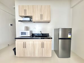 Una cocina compacta con armarios de madera clara, un refrigerador de acero inoxidable, un microondas y una encimera negra.