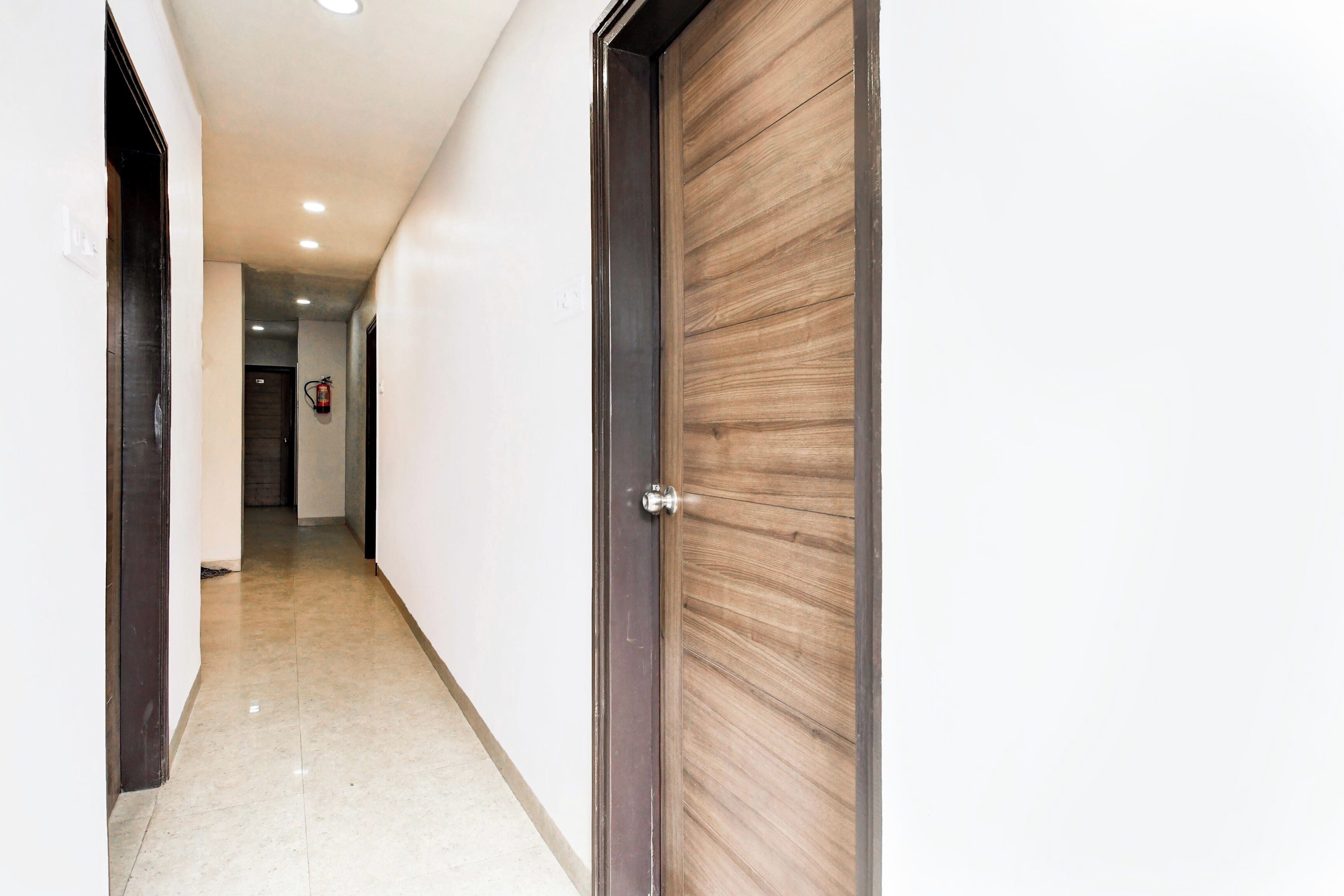 A hallway with a wooden door on the right, a fire extinguisher on the wall, and a door at the end.