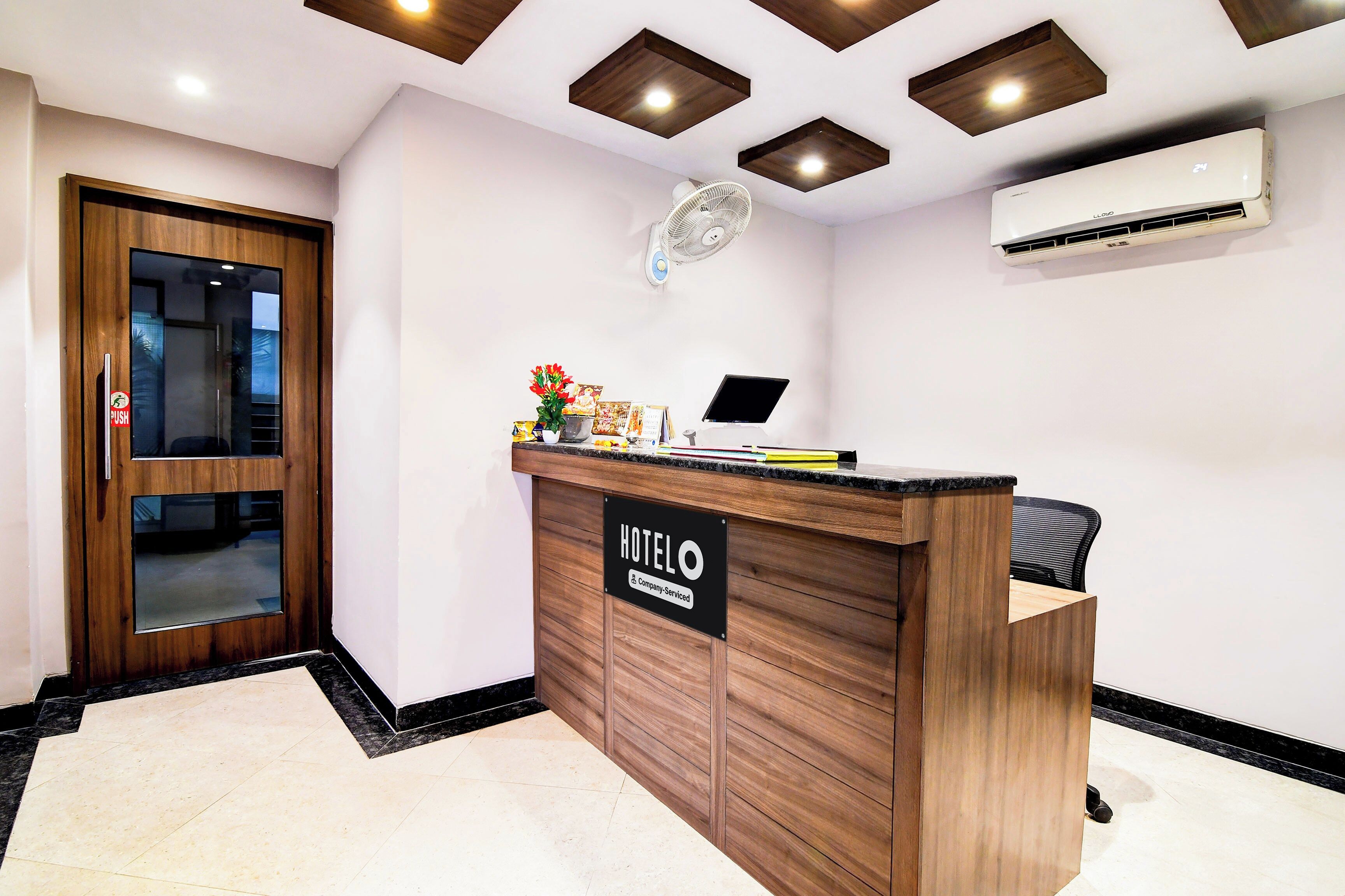 Una recepción de hotel con detalles de madera, un cartel que indica que es un hotel de servicio empresarial, una computadora de escritorio y un aire acondicionado instalado en la pared.