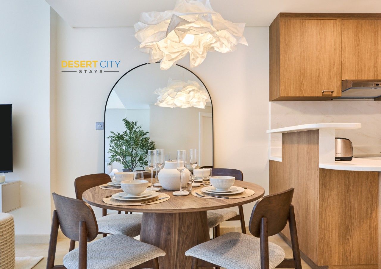 A modern dining area with a round table set for four, a large mirror, and a kitchen area with wooden cabinets.