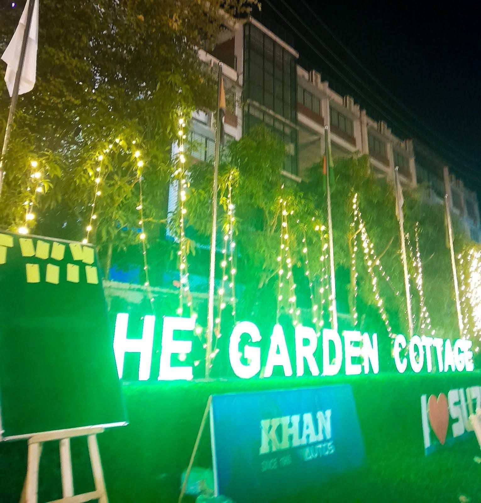 Placa com a inscrição 'HE GARDEN COTTAGE' com luzes decorativas e uma placa com a inscrição 'KHAN' abaixo.