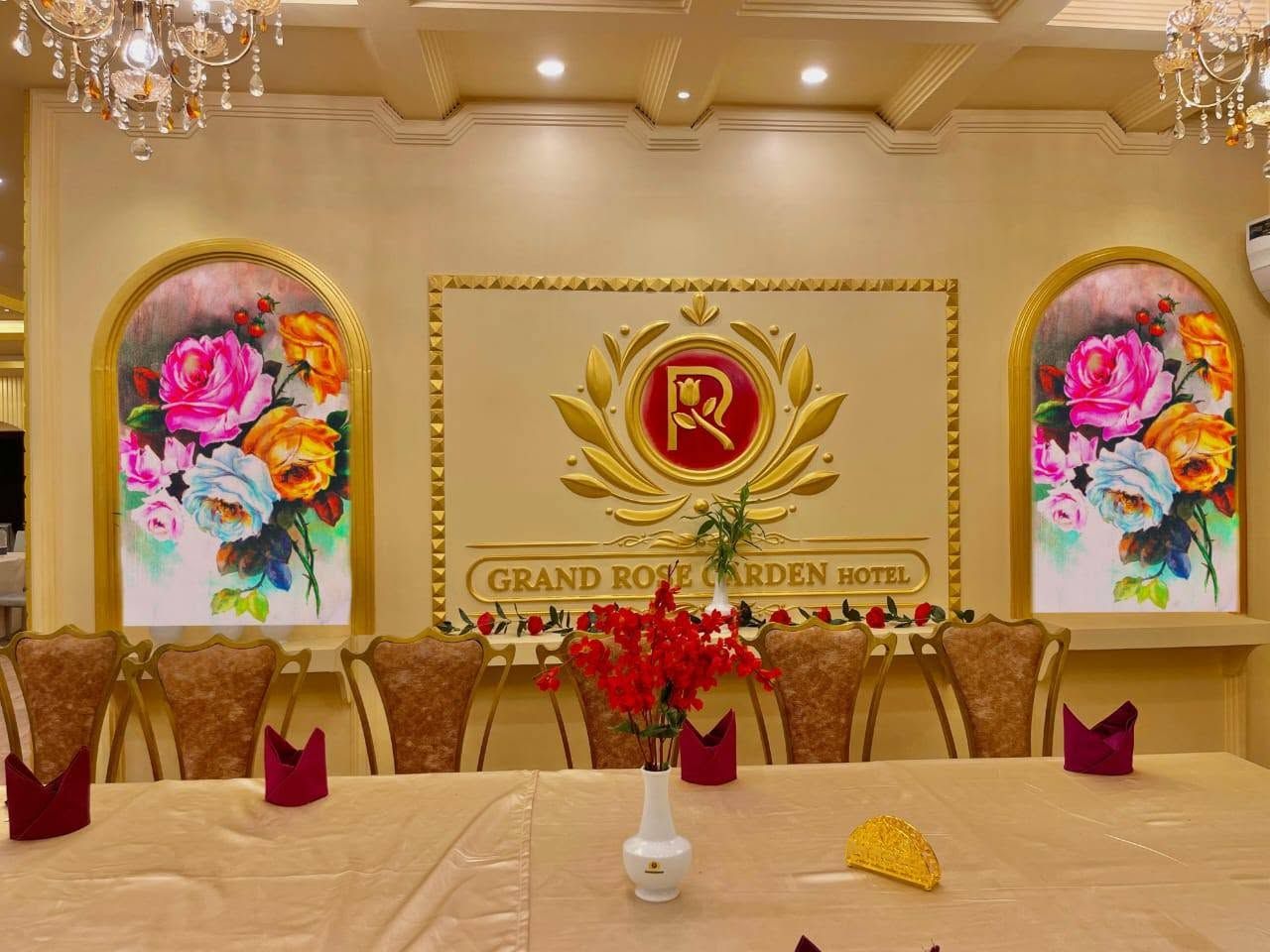 Una mesa de conferencias con un jarrón de flores rojas, un emblema dorado y un cartel que dice 'GRAND ROSE GARDEN HOTEL' en un comedor formal.