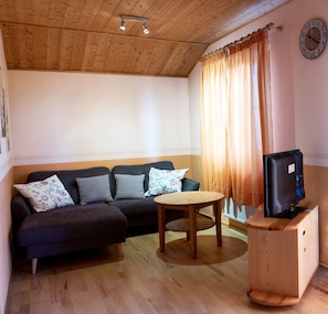 Ein möbliertes Wohnzimmer mit einer Couch, einem kleinen Tisch, einem Fernseher auf einem Holzständer und einer Wanduhr.