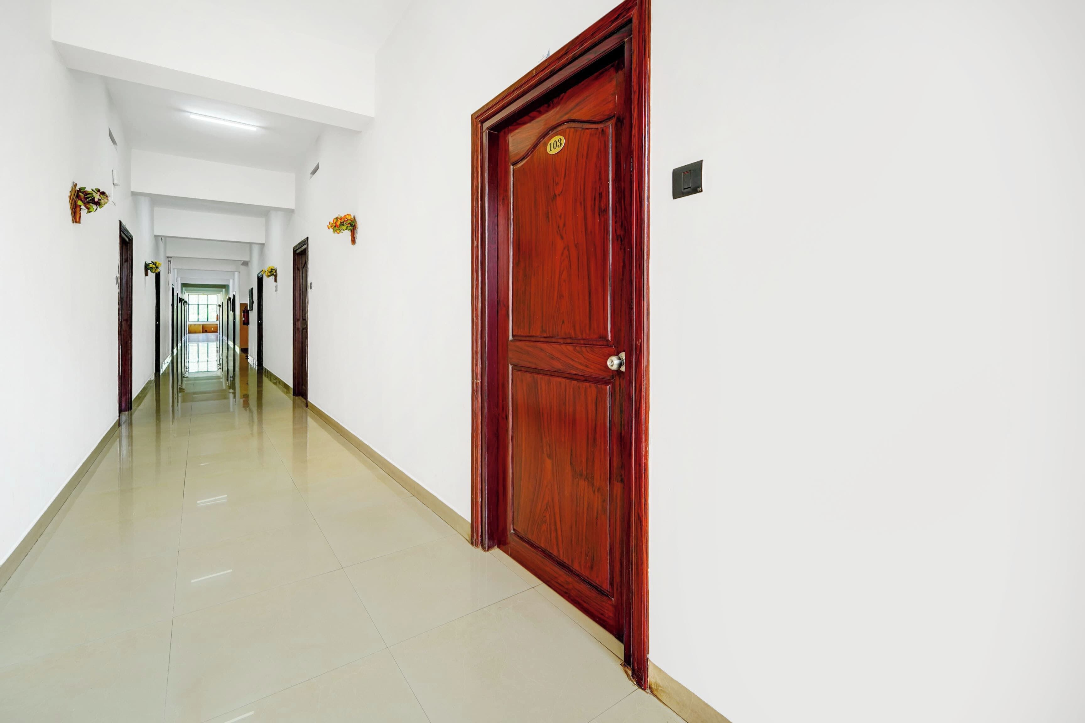Un pasillo largo y estrecho con una puerta de madera a la derecha, paredes blancas y piso de baldosas.