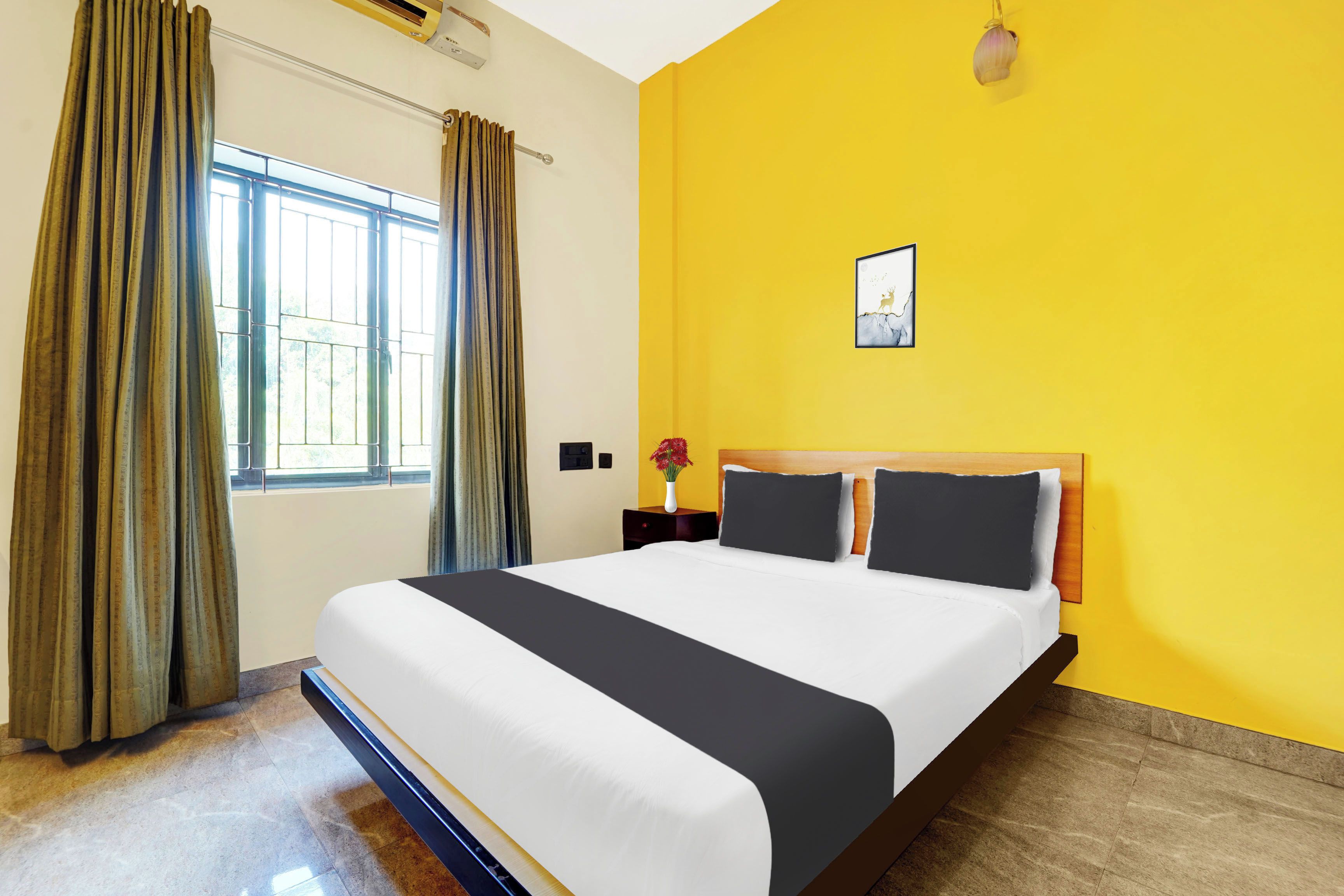 Una habitación de hotel con una cama grande, una ventana con cortinas, una mesita de noche con un jarrón y una pared amarilla.