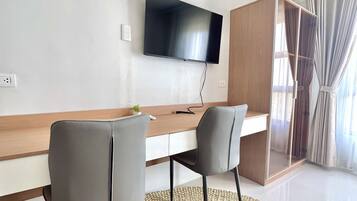 A modern living room with a flat-screen TV mounted on the wall, a wooden desk, two grey chairs, and a woven rug.