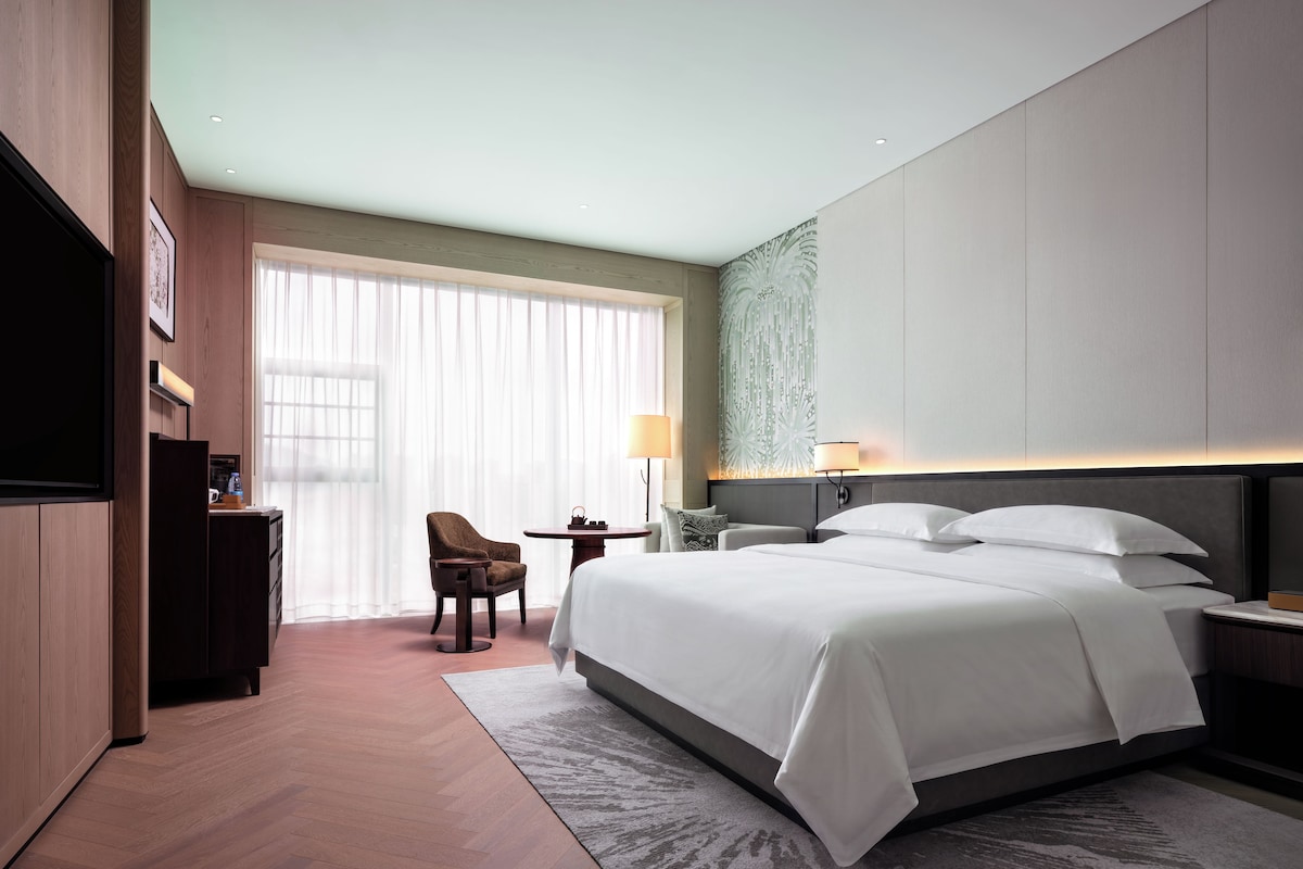 A hotel room with a large bed, a desk with a chair, a TV, and a window with sheer curtains.