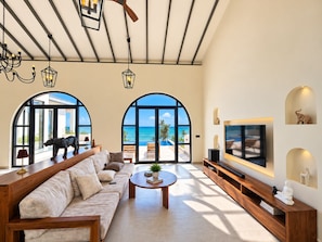A spacious living room with a large sofa, a coffee table, and a TV. There are arched windows offering a view of the ocean.