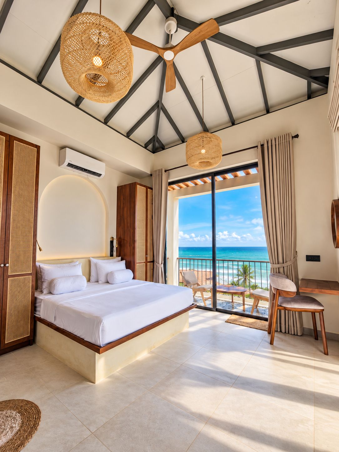 A bedroom with a bed, a ceiling fan, a large window with a view of the ocean, and a balcony with chairs.