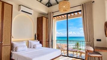 A bedroom with a bed, a ceiling fan, a large window with a view of the ocean, and a balcony with chairs.