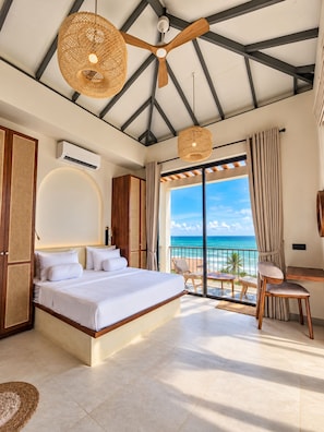 A bedroom with a bed, a ceiling fan, a large window with a view of the ocean, and a balcony with chairs.