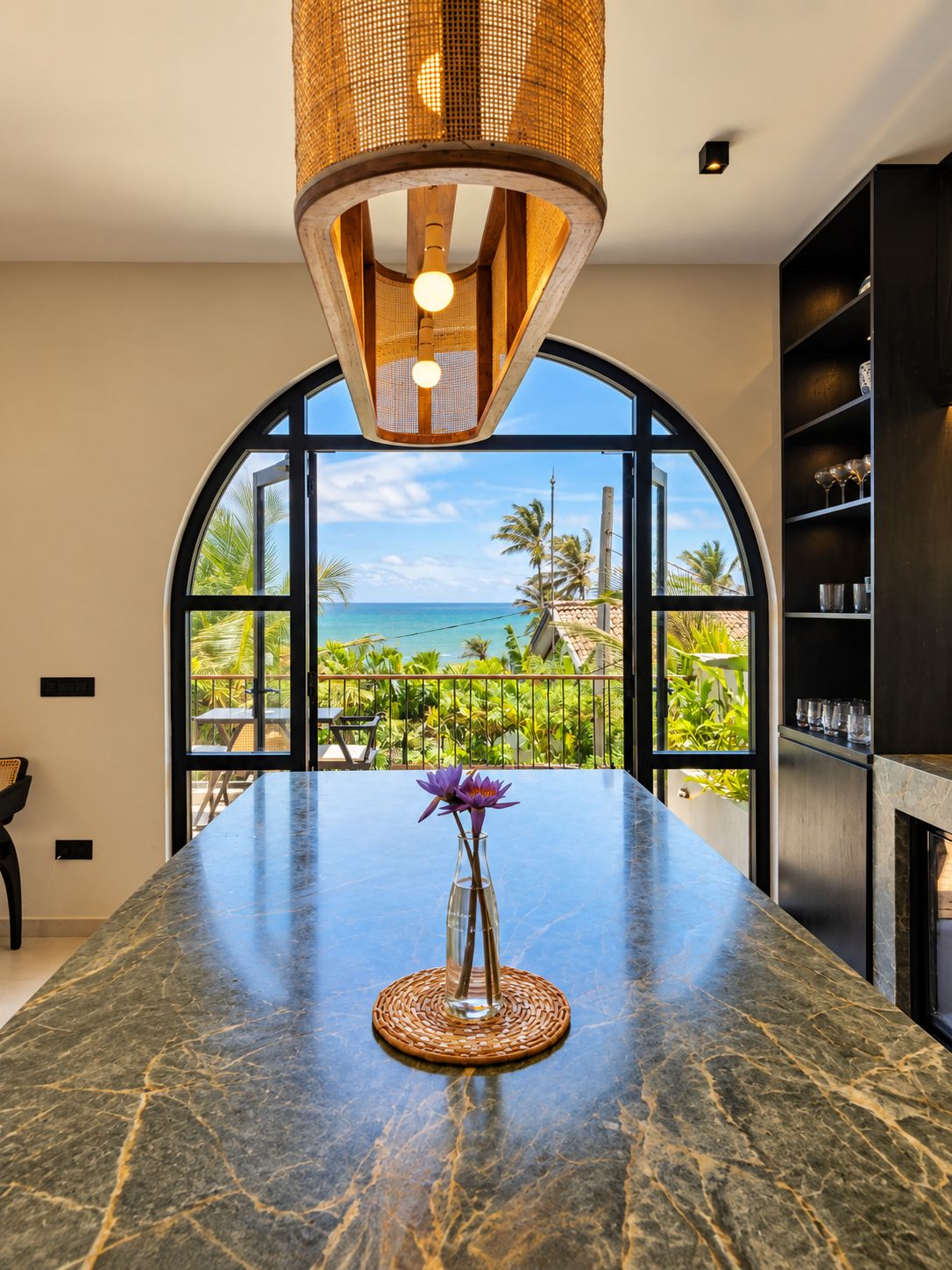 A kitchen with a marble countertop, a vase with flowers, and a view of the ocean through large windows.