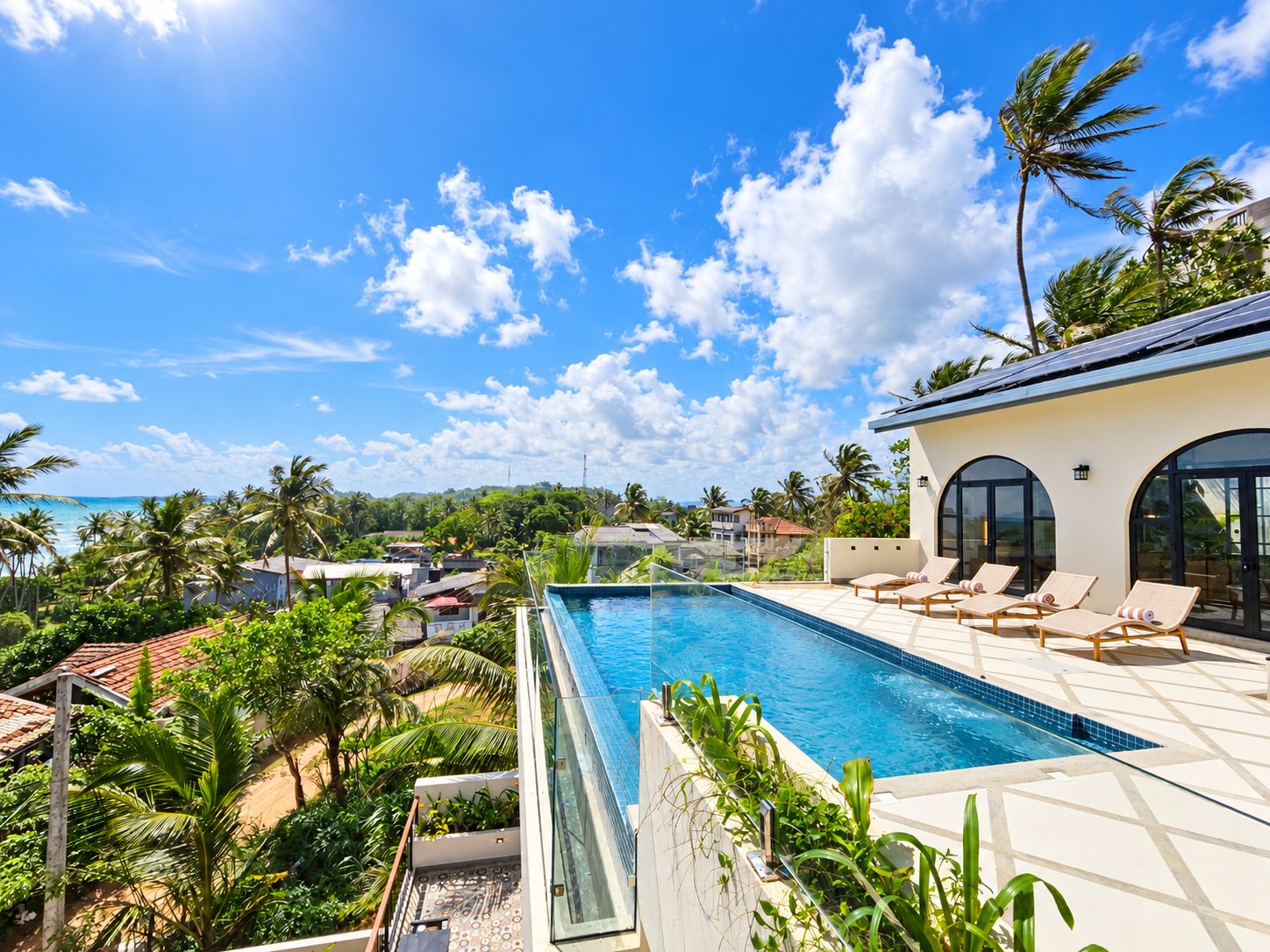 Una piscina infinita en la azotea con sillones de descanso, rodeada de exuberante vegetación tropical y vistas a una zona residencial.
