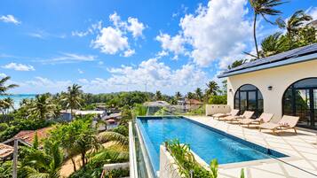 Una piscina infinita en la azotea con sillones de descanso, rodeada de exuberante vegetación tropical y vistas a una zona residencial.
