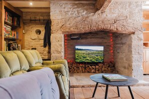 A cozy living room with a stone fireplace, a television, a sofa, and a bookshelf. - Beautiful 4BD Cottage in Minchinhampton (Minchinhampton)