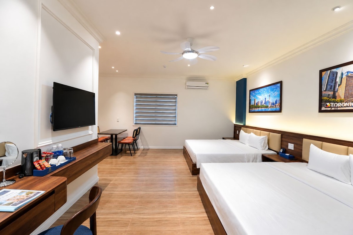 A hotel room with a TV, a desk with amenities, two beds, a ceiling fan, and a window with blinds.