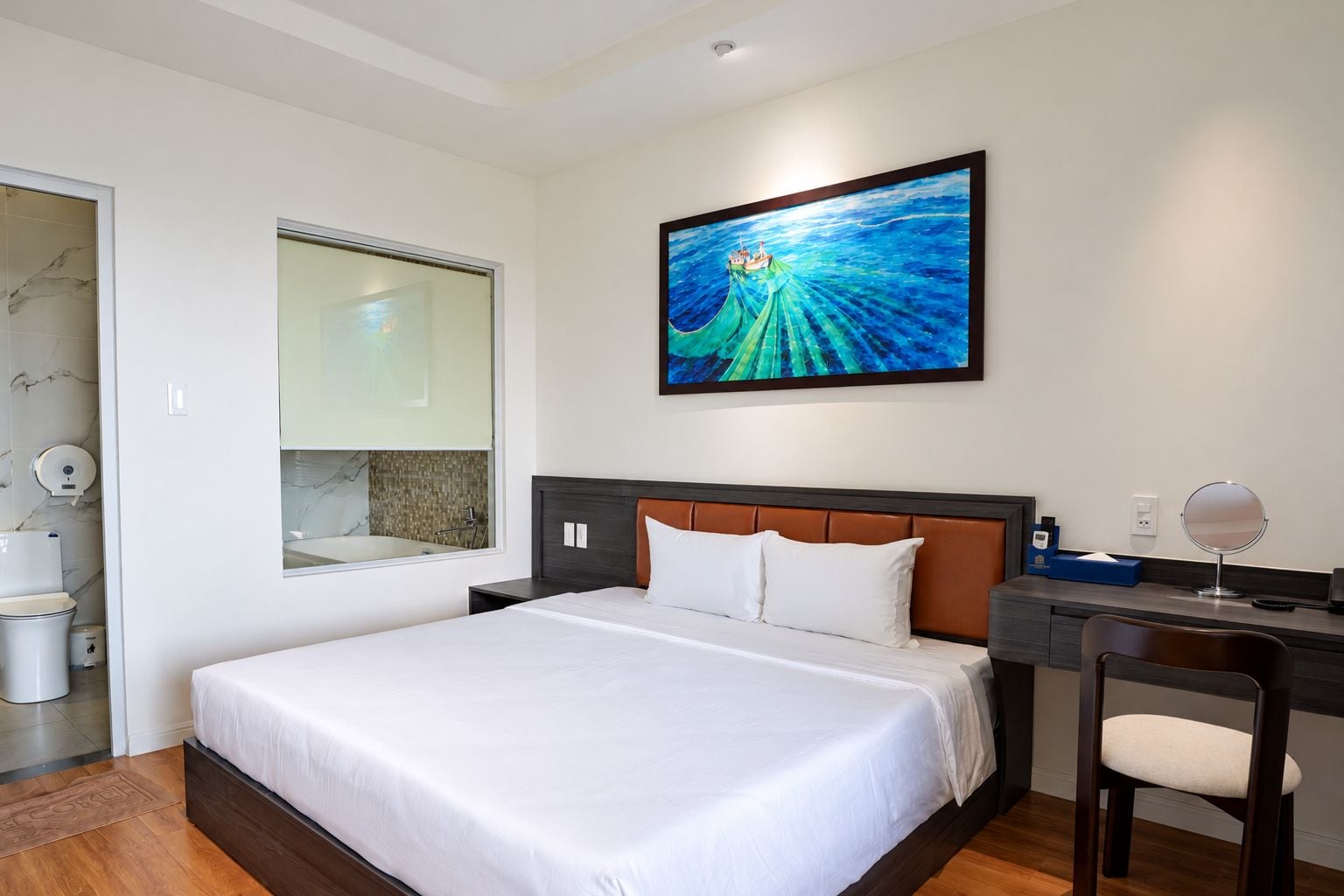 Une chambre d’hôtel avec un grand lit, un bureau avec une chaise, une salle de bain avec un miroir et un tableau au mur.