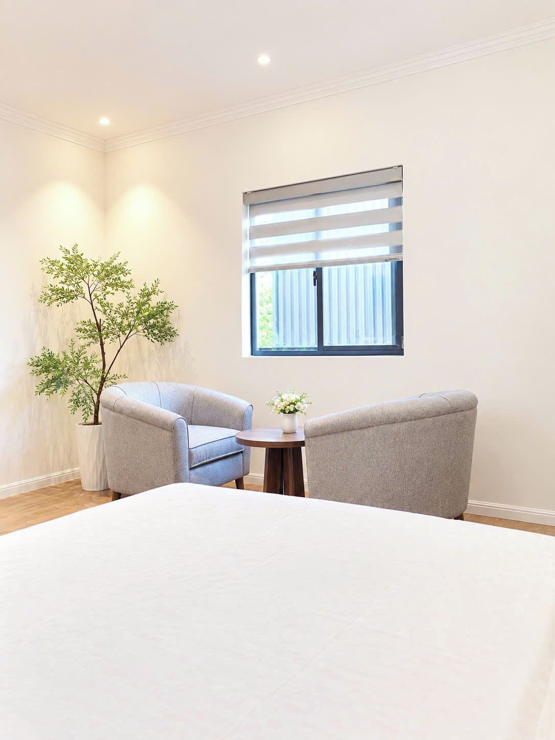 A bedroom with a bed, two armchairs, a small table, a potted plant, and a window with blinds.