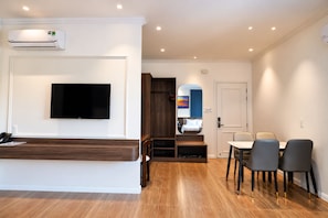 Habitación de hotel con televisión de pantalla plana, aire acondicionado integrado, mostrador de recepción de madera, zona de comedor con mesa y sillas, y un espejo que refleja la habitación.