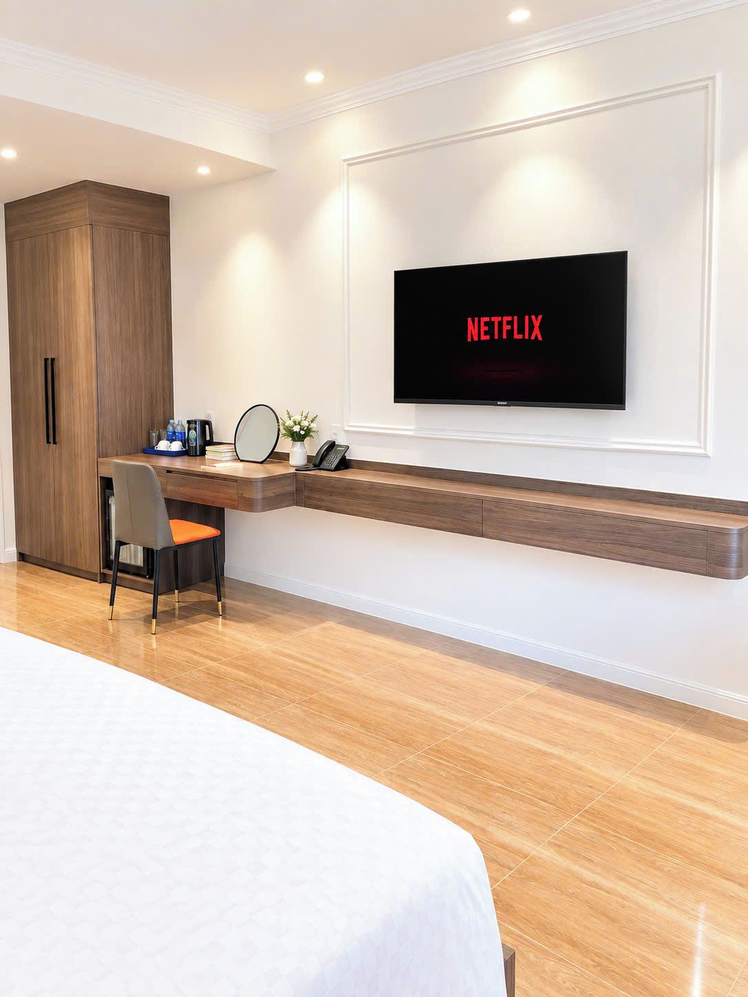 A hotel room with a bed, a desk with a chair, a television mounted on the wall, and a wooden wardrobe.