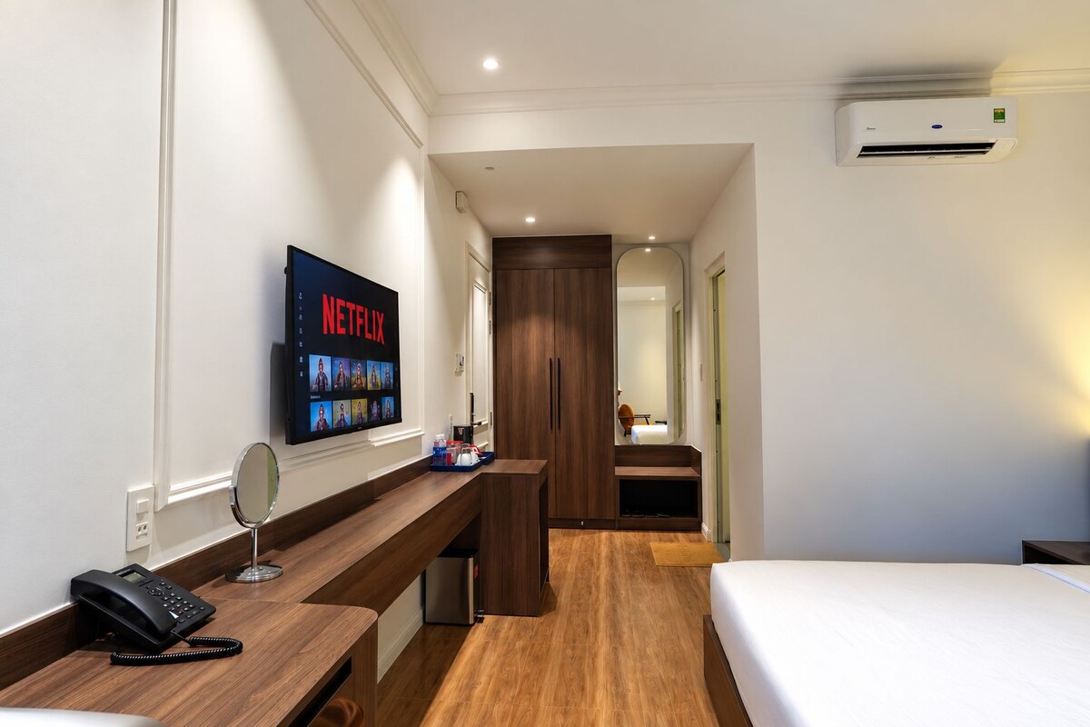 A hotel room with a bed, a desk with a phone, a wall-mounted TV displaying Netflix, and a hallway leading to another room.