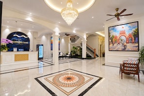 A hotel lobby with a reception desk, a chandelier, a staircase, and a large painting.