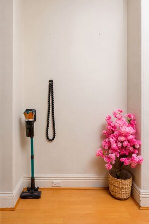 Eine Ecke eines Zimmers mit einem Staubsauger, einem schwarzen, verzierten Gegenstand an der Wand und einem Korb mit rosa Blumen.