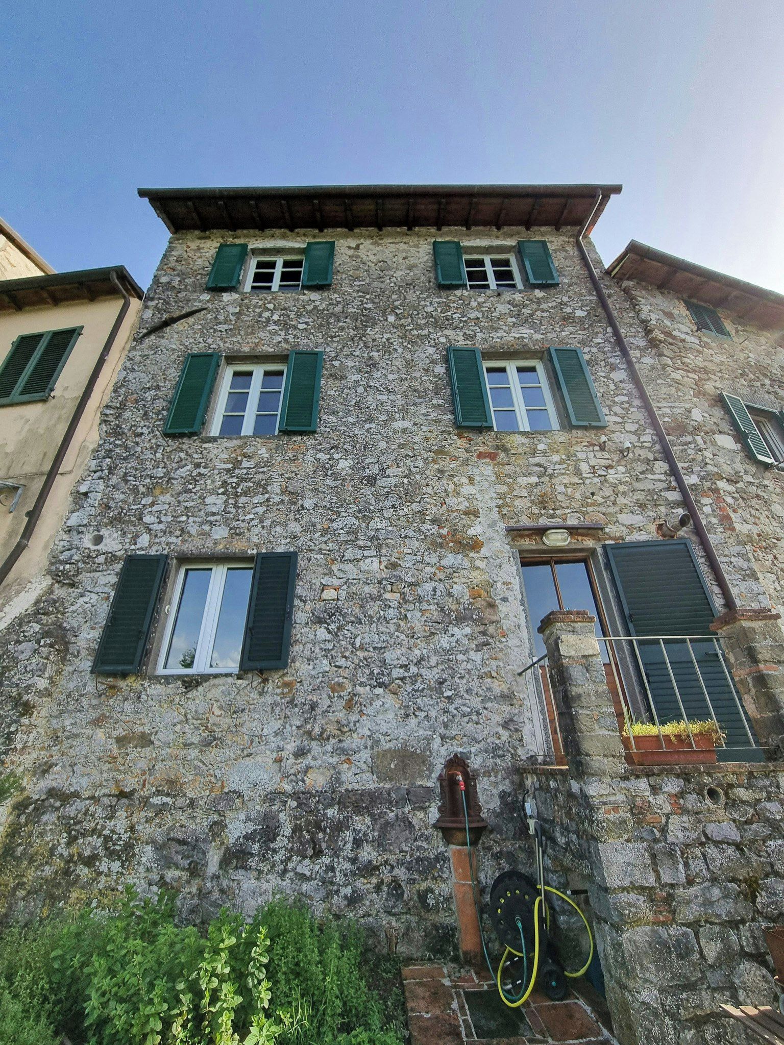 Un edificio in pietra su più piani, con persiane verdi, un rubinetto a vista e un portabici.