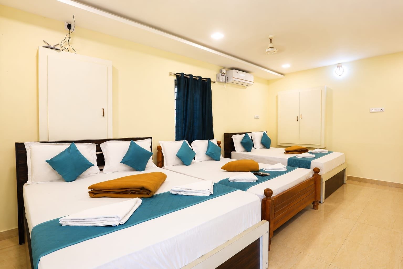 A hotel room with three beds, each with a white mattress and teal bedspreads. There are wooden headboards, and the room has a tiled floor.