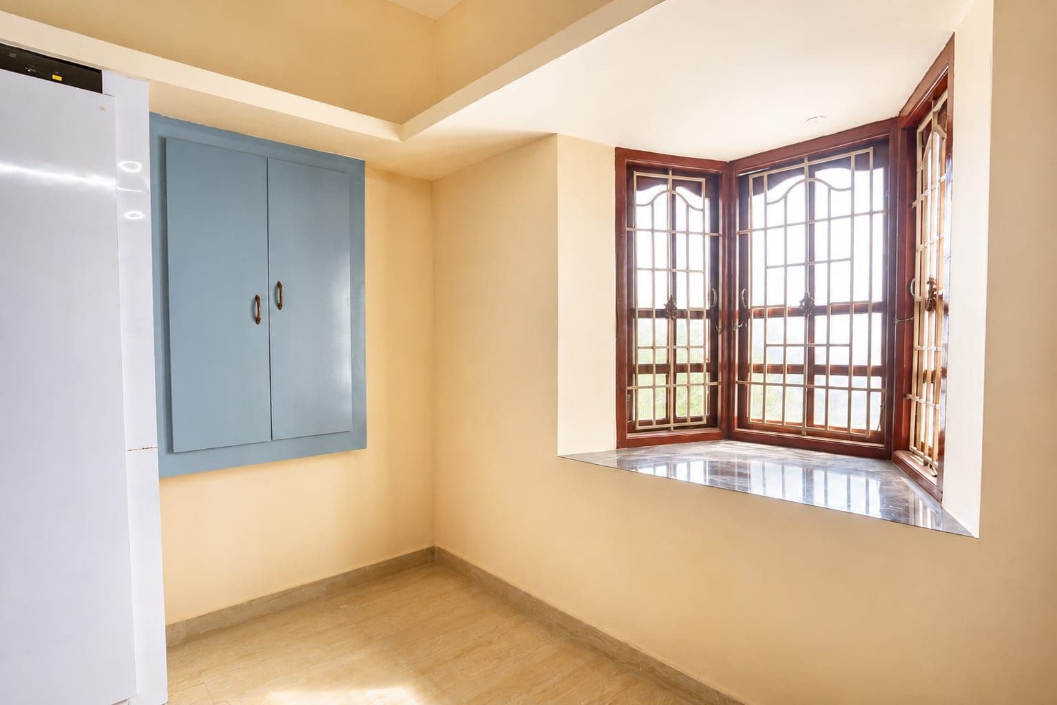 A room with a blue wardrobe, a corner window with wooden frames, and a white refrigerator.